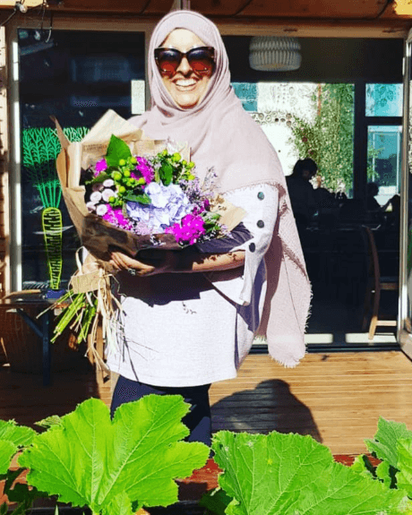 A lady stands wearing a head scarf and large sunglasses. She is holding a colourful bouquet of flowers