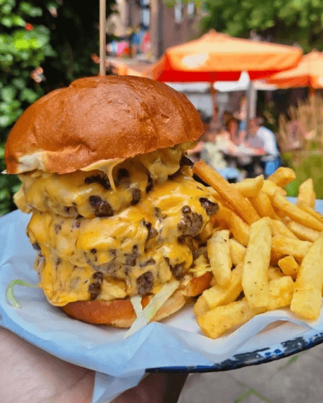Three beef burger patties piled on eachother, dripping with cheese and served in a brioche bun with fries. The plate is held up outside in a seating area with large orange umbrellas.