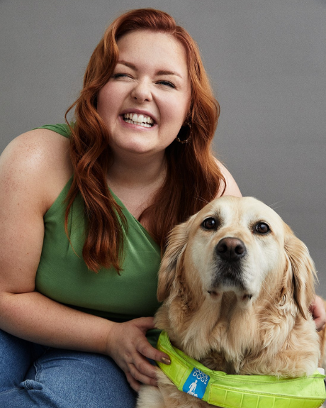A lady with red, waved hair smiles at the camera whilst holding onto a sandy coloured, golden retriever who is her guide dog.