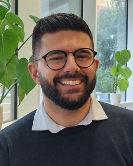 A man with well groomed, dark hair smiles at the camera form behind, black, round lense glasses. He has a short beard and is wearing a white shirt, with a navy blue jumper over.
