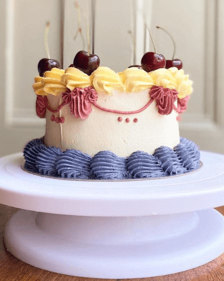A cream cake with lilac, pink and cream piping detail topped with cherries