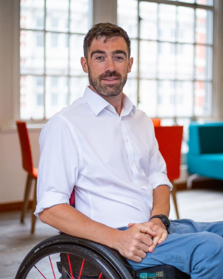 A man smiles at the camera. He is wearing a white shirt and blue trousers. He has short, dark hair and stubble. He is sitting in a black wheelchair, in a large open room with tall windows behind him.