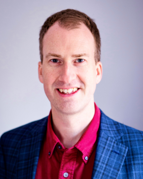 A man wearing a red shirt and blue, tartan suit jacket smiles at the camera.