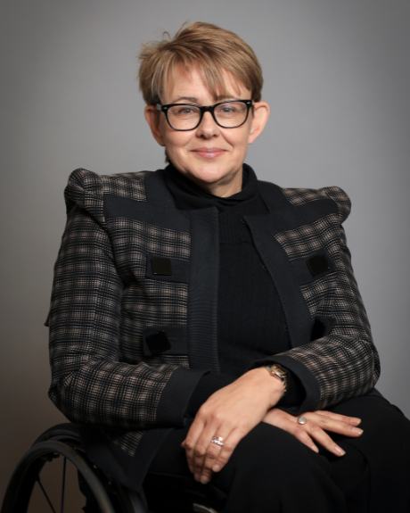 A lady wearing a black outfit including a jacket with prominent shoulder pads, smiles at the camera. She has short brown hair with highlights and black, thick framed glasses. She is sitting in a wheelchair.