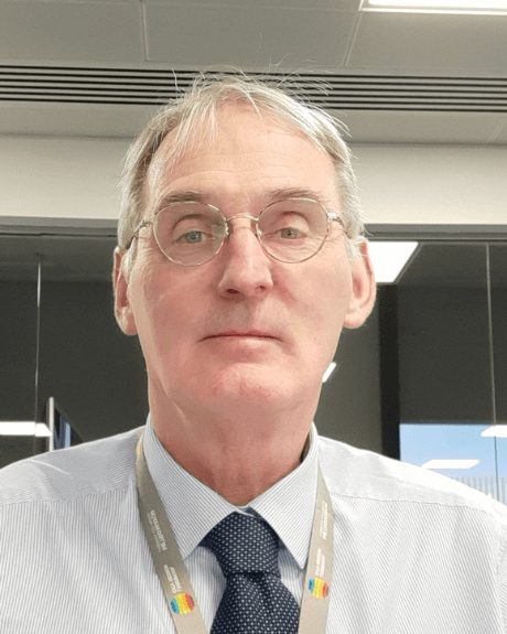 A man looking at the camera wearing a shirt and navy blue tie. He has a lanyard round his neck and is wearing glasses.