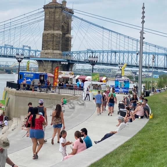 People mingle at a festival. In the background are several food trucks in front of the Roebling Suspension Bridge.