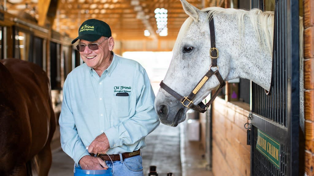 An elderly man smiles while feeding a white horse in a barn at Old Friends Thoroughbred Retirement Farm..