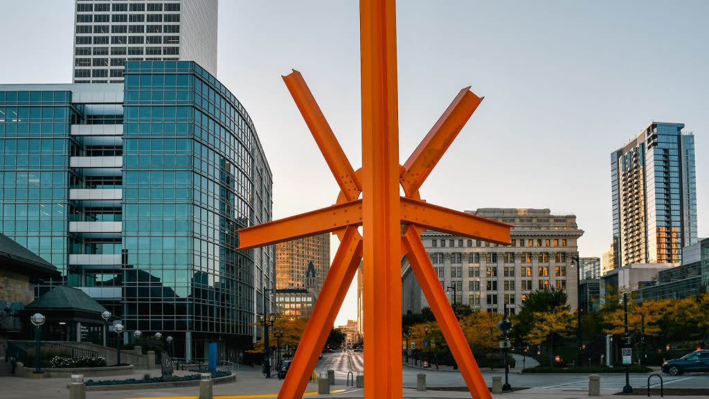 An orange steel sculpture titled The Calling stands at the center of Milwaukee’s O’Donnell Park, with modern glass buildings and historic architecture framing the background. The artwork's bold, angular beams stretch upward and outward, glowing in the light of the setting sun. The streets surrounding the sculpture are quiet, with a few pedestrians and autumn trees lining the sidewalks.