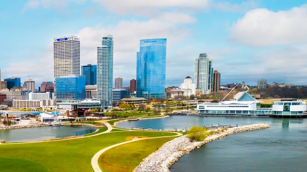 A bright daytime view of Milwaukee’s lakefront featuring modern high-rise buildings, the Milwaukee Art Museum with its iconic white wings, and curving walking paths along the shoreline. Lake Michigan’s blue water frames the foreground while the downtown skyline rises behind the green park space.