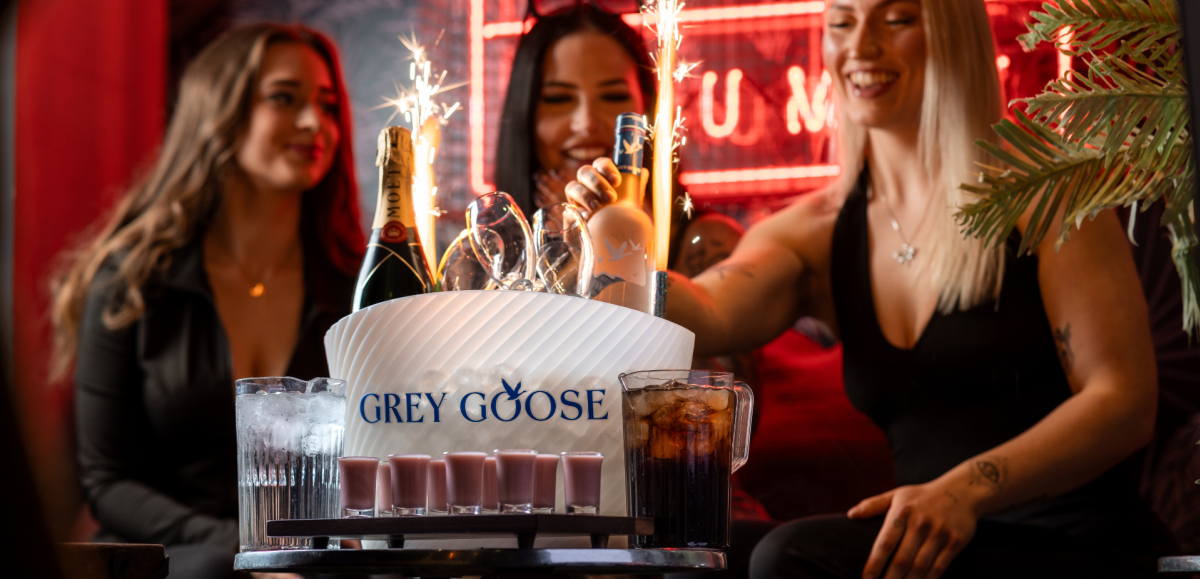Three women celebrating at a nightclub table with a Grey Goose ice bucket holding champagne and vodka bottles, sparklers lit, surrounded by glasses and drinks."