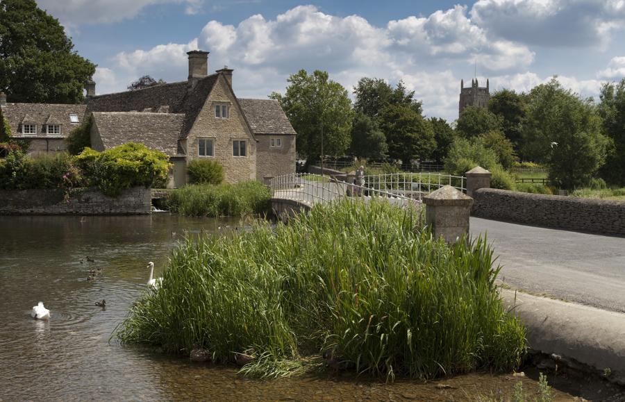 Swans and ducks swim on the river by a bridge and a Cotswold stone house in Fairford