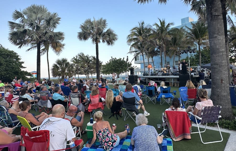 A crowd of people sitting in lawn chairs and on towels, on the Pompano Beach Great Lawn at dusk for Music Under the Stars
