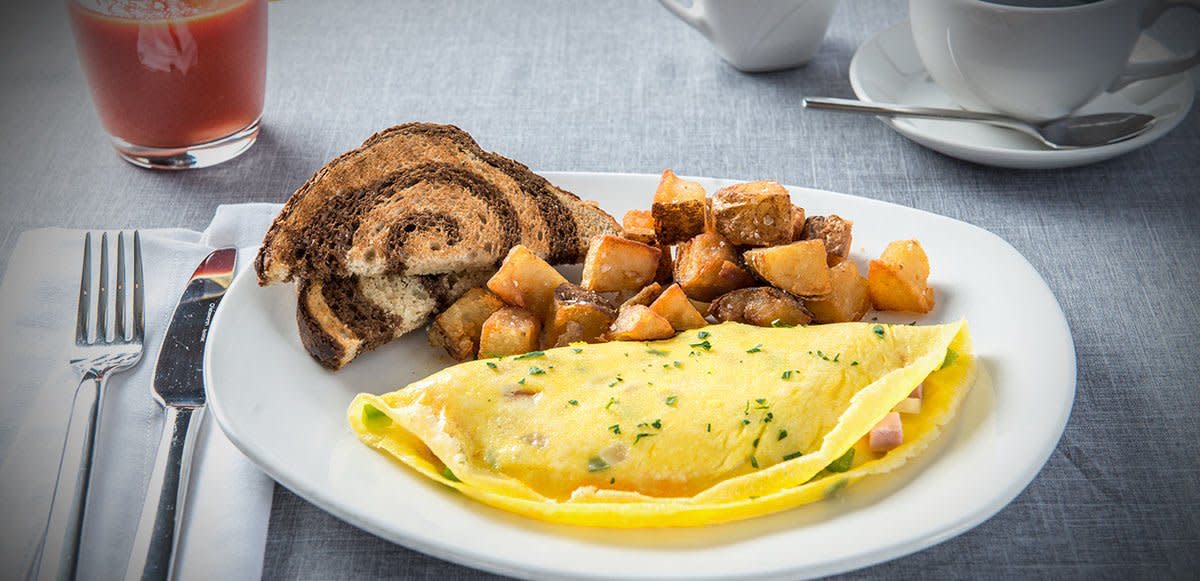 A classic breakfast plate featuring a folded omelet garnished with chopped herbs, served with golden-brown breakfast potatoes and two slices of marble rye toast. The meal is presented on a white plate with a glass of juice, a cup of coffee, and silverware on a light gray tablecloth.