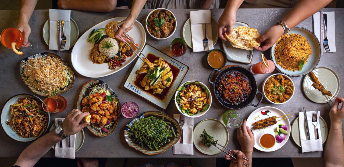 The camera shows an overhead view of a table at Phat Eatery. It features more than a dozens plates, cups, bowls, and skillets piled high with various Asian dishes: orange chicken, grilled fish, rice pilaf, pita bread, kebobs, etc. At least six people are seated at the table, but only their hands are visible as they reach for their glass, use chopsticks, or gently tear the food apart with their fingers.