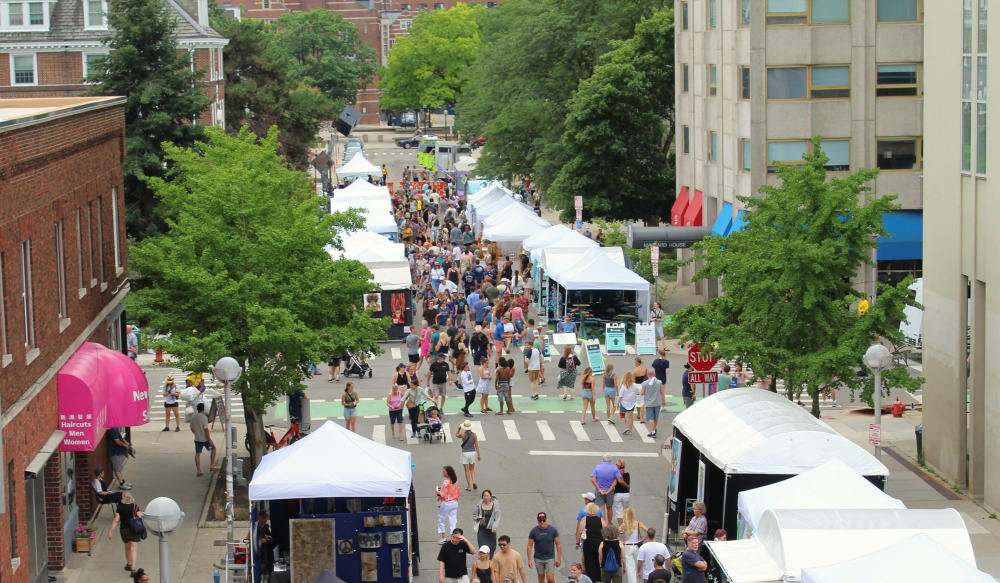 bird's eye view of a street lined on both sides with white vendor tents. Pedestrians walk the streets as they browse the tents.