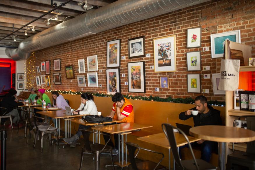 row of small tables with people sitting, drinking coffee and looking at phones or laptops
