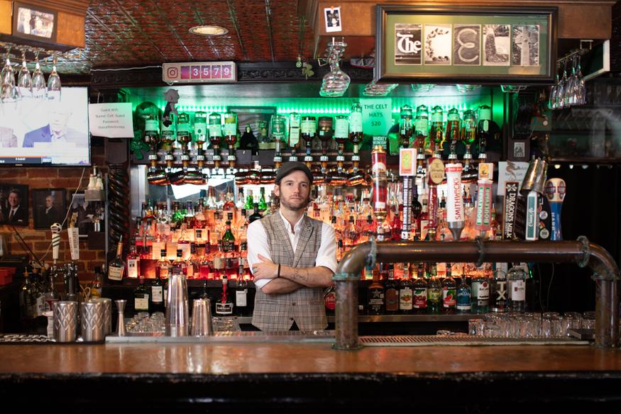 Man in a vest and driving hat stands, arms folded, behind a fully-stocked bar