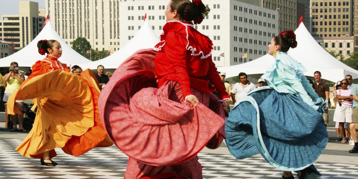 Women dancing in traditional, colorful dresses on stage in front of skyline at Festival Latino