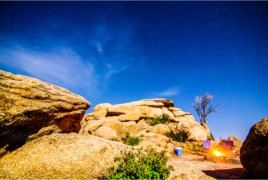 Starry night sky over Vedauwoo, with a campfire glowing between large rock formations and camping chairs visible, evoking a sense of adventure and tranquility.
