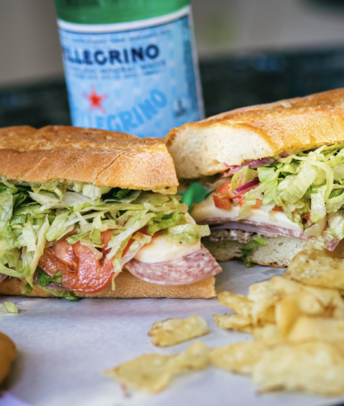 Italian sub sandwich with sliced meats, cheese, tomatoes, shredded lettuce, red onion, and dressing, served with potato chips.