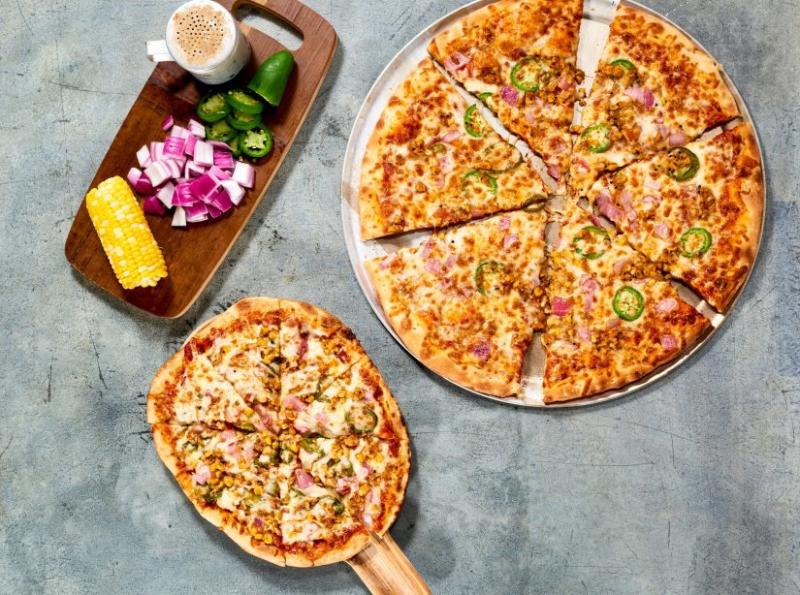 An oversized pizza on a metal tray next to a small pizza on a pizza peel and a wooden tray with corn on the cob, sliced onions and jalapenos from Pizza Karma