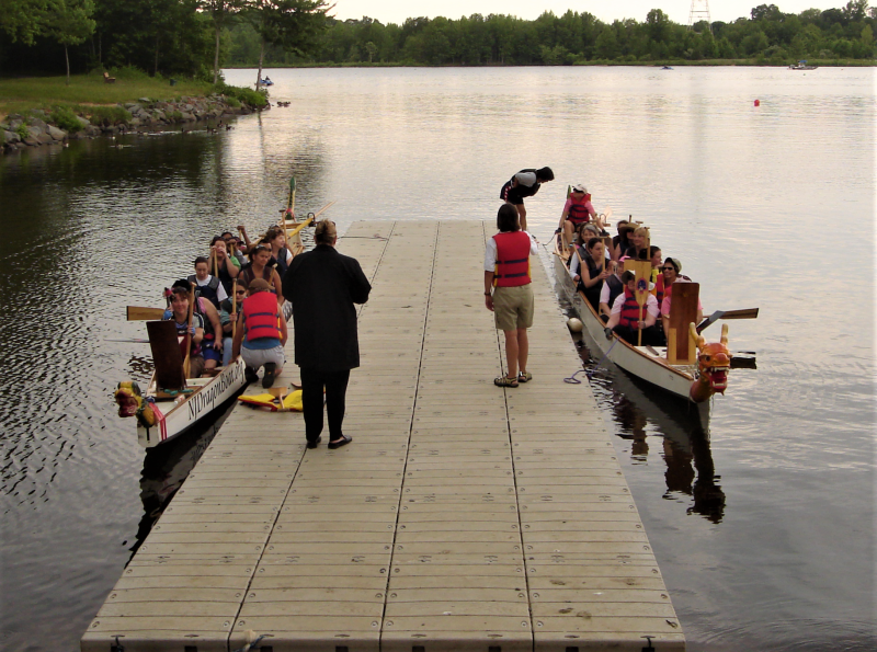 Two boats float along the side of wooden dock.Children in life jackets sit inside the boats holding oars.