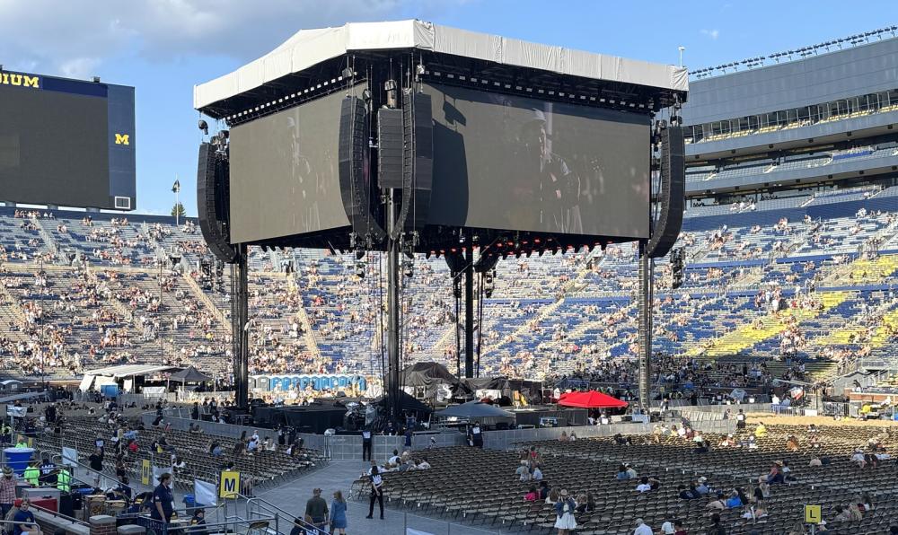 Inside the michigan stadium with a concert set-up on the field. The jumbotron stands tall in the middle of the field with two screens visible.