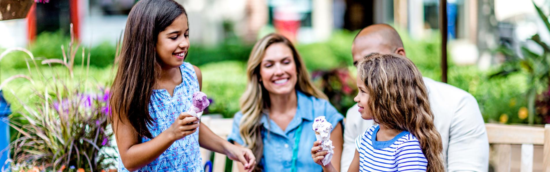 A mom and dad sit on a bench while happily watching their two girls hold ice cream cones