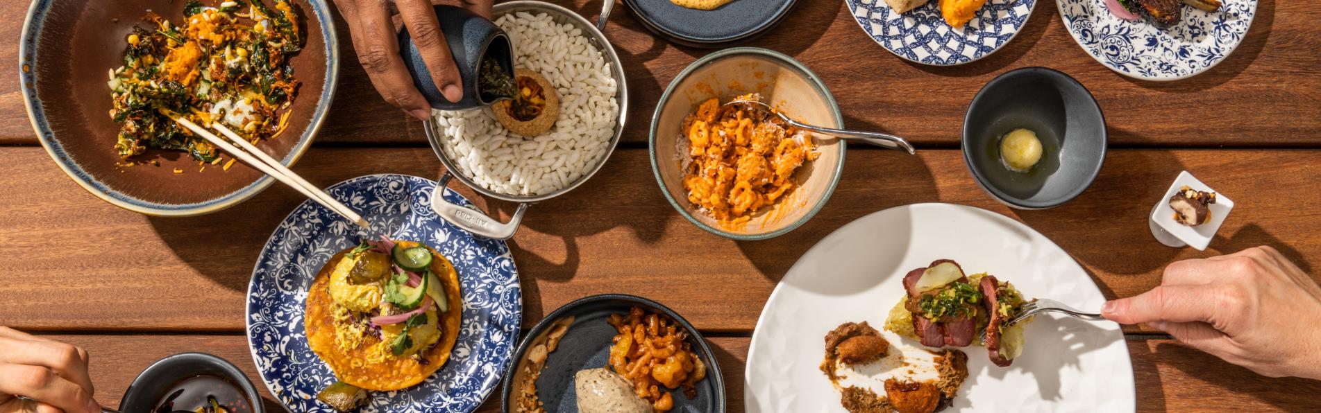Several plates of food on a wooden table with hands reaching out to pour sauce, grab with forks and pull bread apart