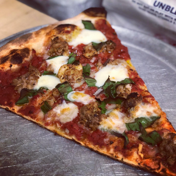A large slice of pizza topped with chunks of sausage, melted mozzarella, tomato sauce, and fresh basil, served on a metal pizza pan.