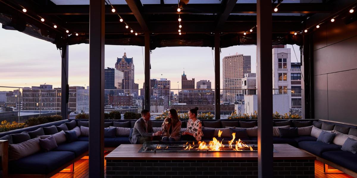Three people sitting around a rooftop fire pit at sunset, enjoying drinks with a view of downtown Milwaukee’s skyline at The Outsider rooftop bar.