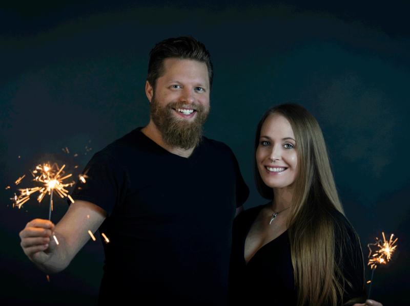 Chris and Bethany Ahern stand smililng with sparklers in their hands. Dark backdrop