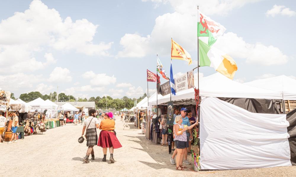 couple walking on dirt grounds, dressed in kilts between lined up vendors in white tents selling goods related to the Michigan Celtic Festival. Blue skies with white clouds, trees in distant background.