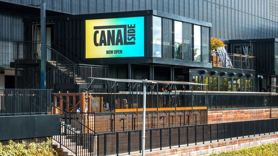 Canalside at Co-op Live in Manchester, showing a modern building with large Canal Street signage, metal railings, brick walkways and greenery alongside the canal.