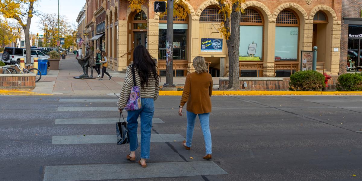 Two women walk across a crosswalk in an autumn urban setting, surrounded by historic brick buildings and colorful trees. They appear relaxed.