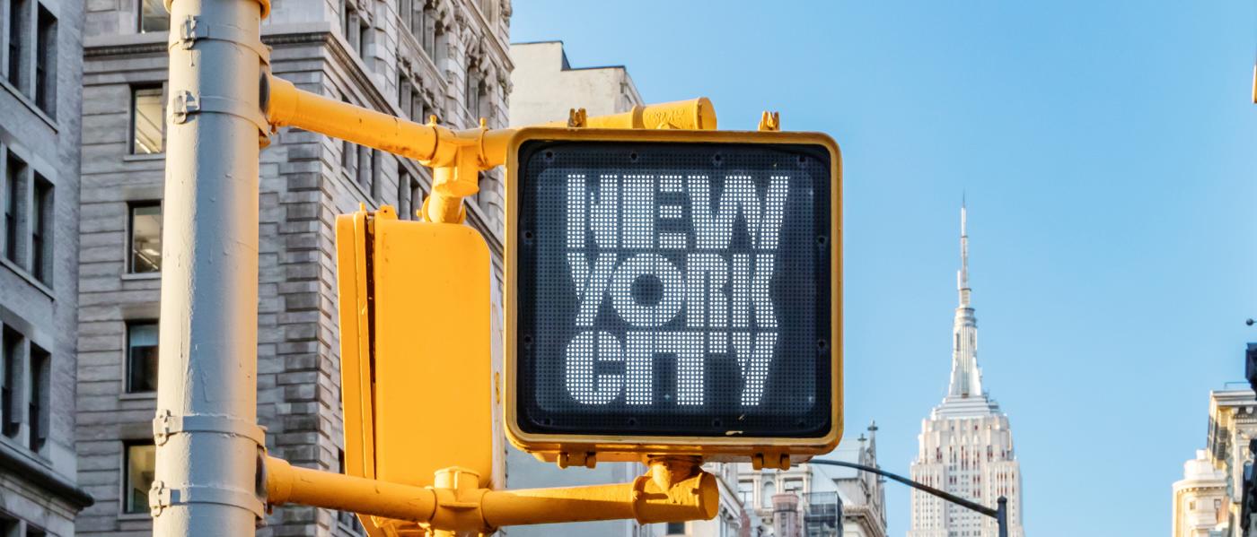 A street-side walk sign, with the Empire State Building in the background,  displaying the logo for New York City Tourism + Conventions