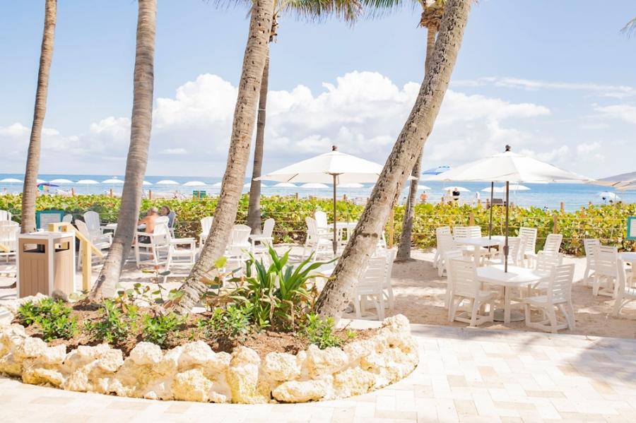 Several tables and chairs with umbrella set in the sand with an ocean view, for table service.