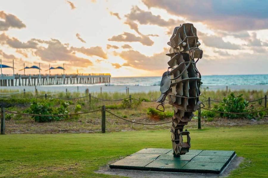 A sculpture of a seahorse sitting beachside on a patch of green grass with the Fisher Family Pier in the background at sunrise in Pompano Beach.