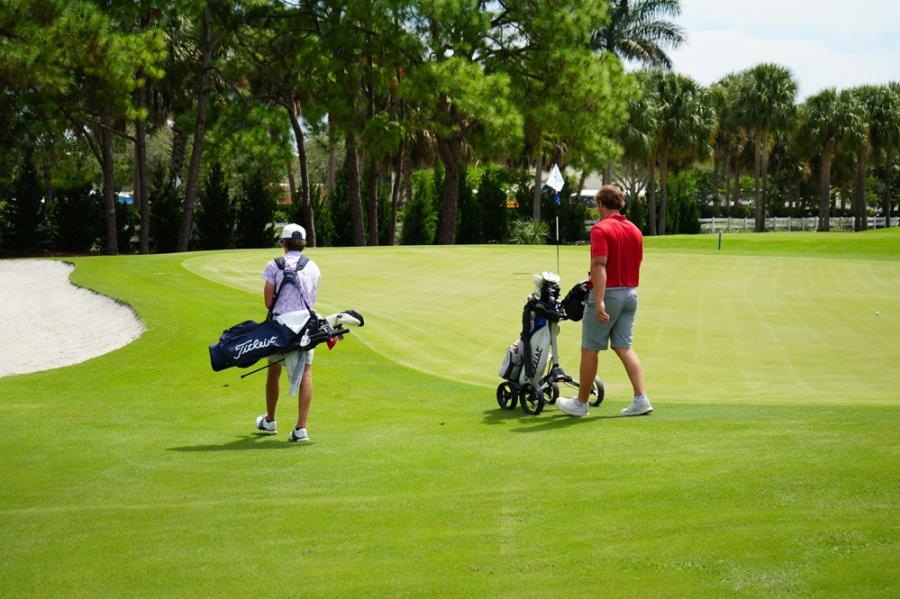 A father and son walking on a beautiful, green golf course in Pompano Beach. Dad's bag is on a push cart, and the son is carrying his bag.