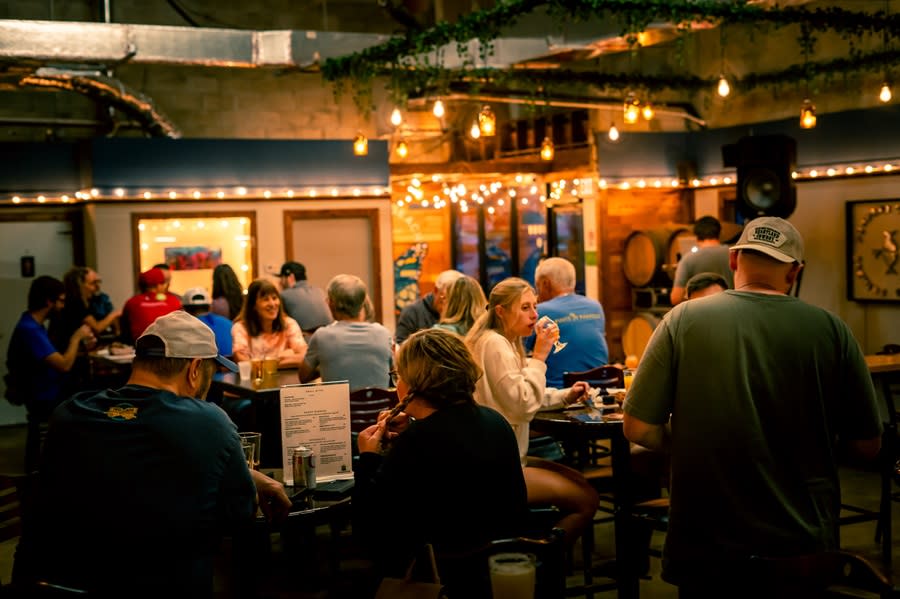 A cozy brew pub with bistro lights hanging around the perimeter, filled with couples and small groups dining and enjoying glasses of beer. One couple is reading a menu.