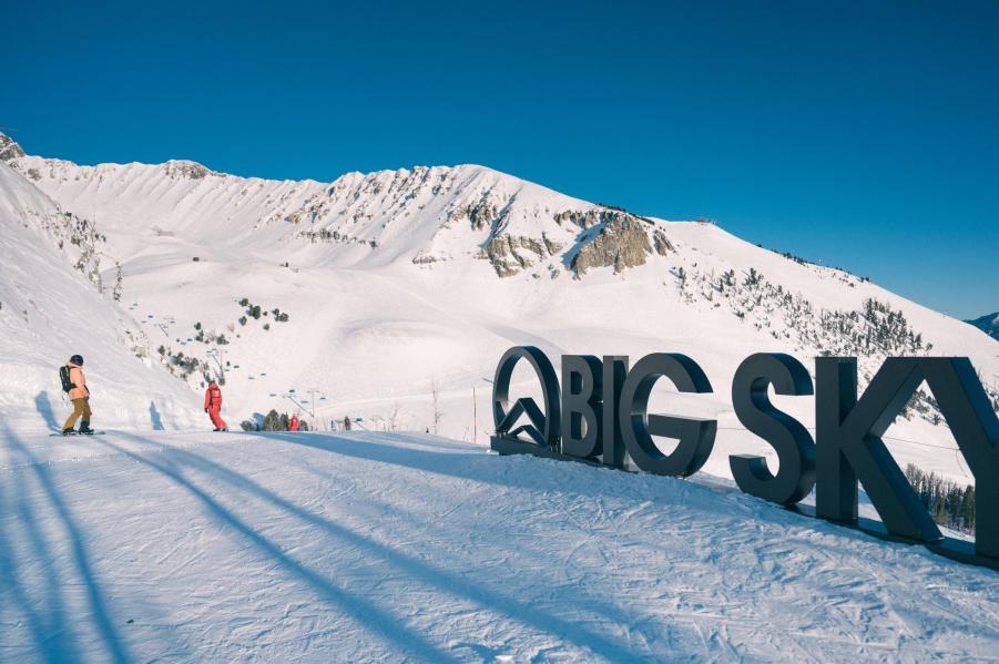 Snow-covered mountains under a clear blue sky, with skiers near a large "BIG SKY" sign. The landscape features rolling slopes and distant peaks.