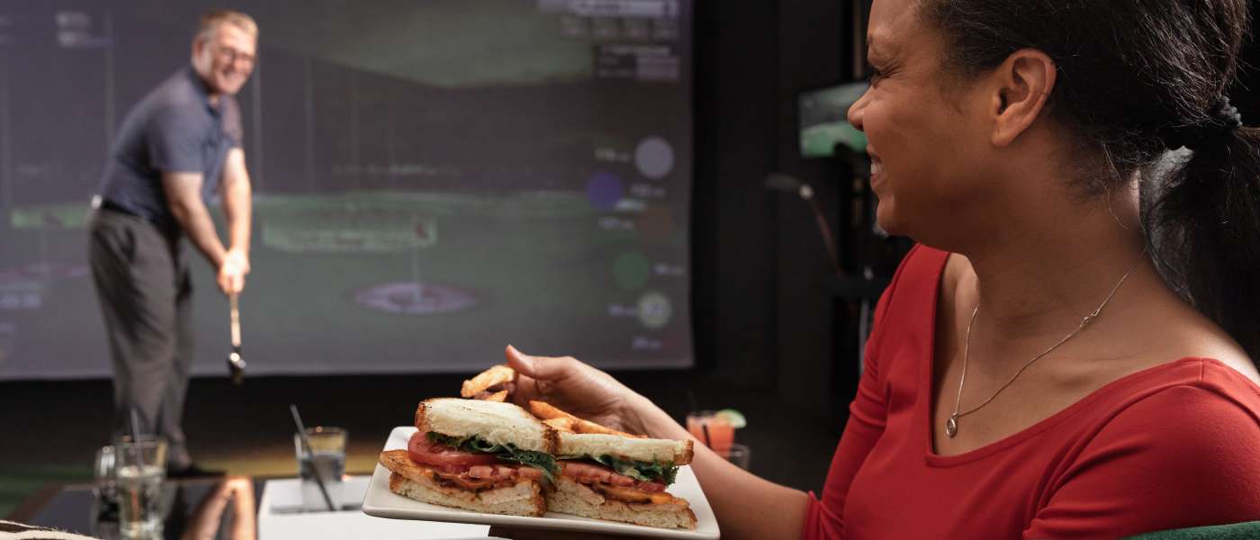 Woman enjoys sandwich while watching her husband play a virtual game of golf at the Main Course