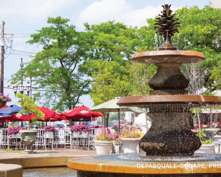 The fountain in Depasquale Plaza surrounded by outdoor dining tables and bright red umbrellas.