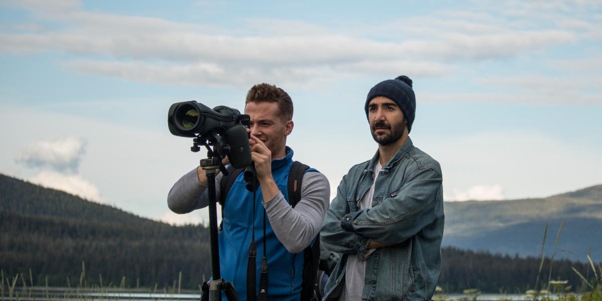 A couple of photographers standing behind their camera with tree-filled mountains behind them in Juneau, AK.