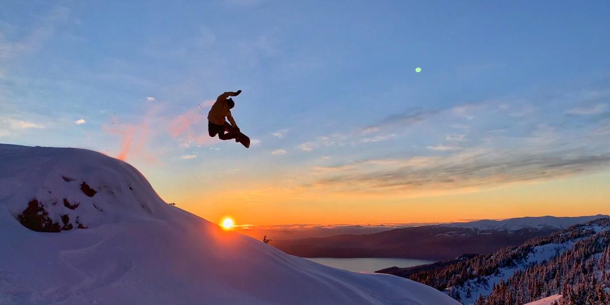 A snowboarder hitting a trick jump mid-air off the Eaglecrest mountain in Juneau as the sun sets over the icy blue waters.