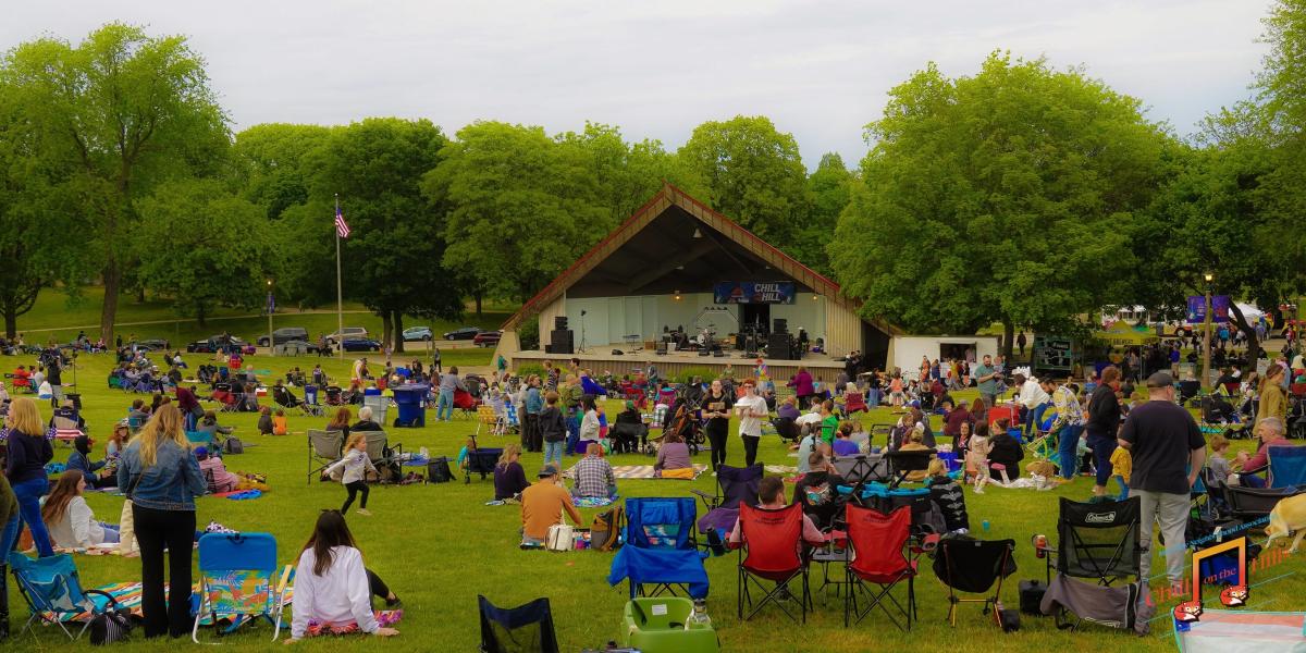 A large crowd gathers on the hillside at Chill on the Hill in Humboldt Park, relaxing on lawn chairs and blankets while facing the outdoor bandshell stage. Families and groups spread across the grassy lawn as the stage is set for live music, surrounded by mature trees on an overcast evening.