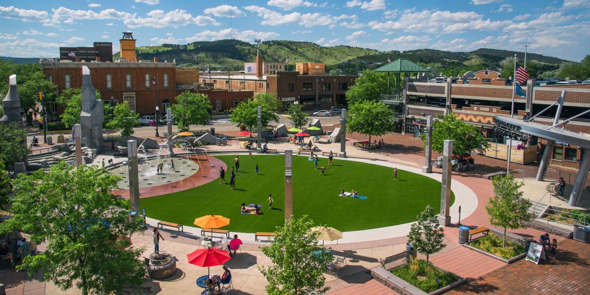 Main Street Square with lush green grass, people relaxing, kids playing near fountains, umbrellas providing shade, and historic brick buildings surrounding.