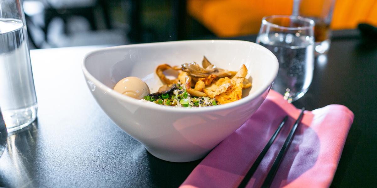 A bowl of ramen on a dark table, featuring a soft-boiled egg, mushrooms, and green onions. Nearby, there is a glass of water and black chopsticks on a pink napkin.