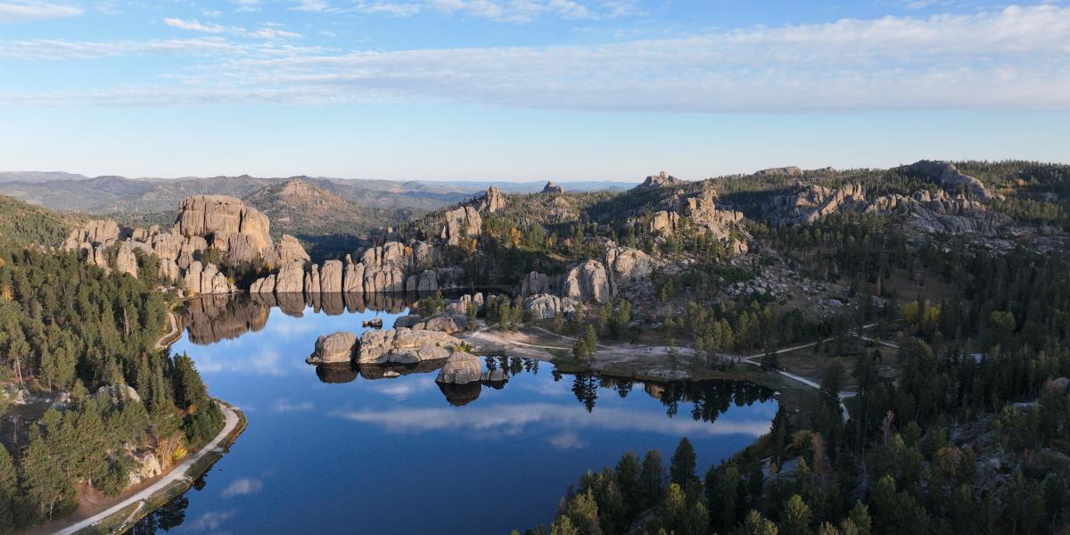 A serene lake reflects rugged rock formations and dense pine forests under a blue sky, evoking tranquility and natural beauty.