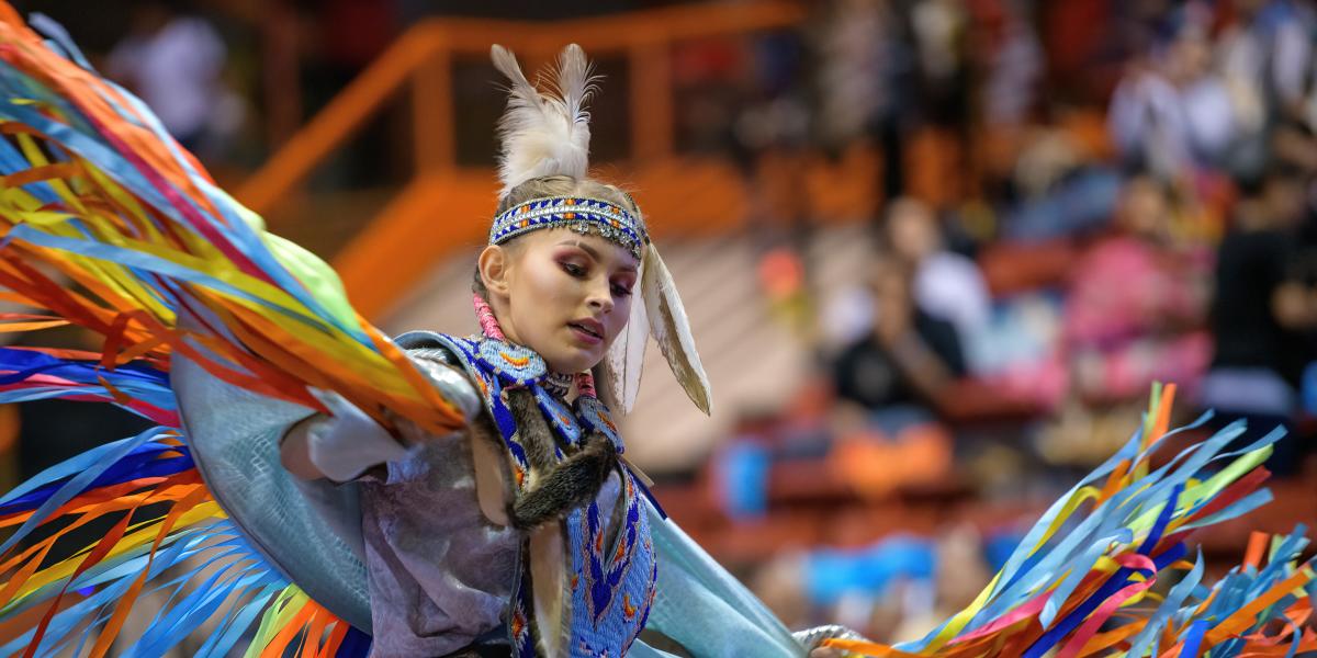 A dancer in vibrant traditional attire performs dynamically. She wears a feathered headdress and colorful ribbons. The atmosphere is lively and festive.